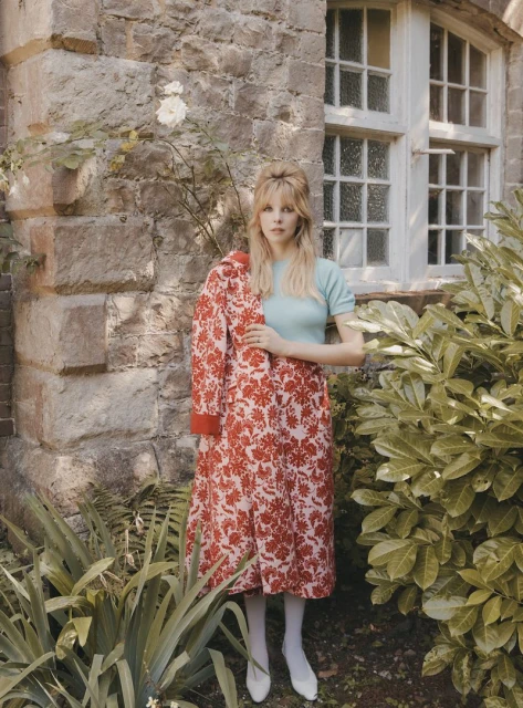 Blonde woman in floral dress poses in garden setting in front of stone building, holding the dress jacket on her shoulder.