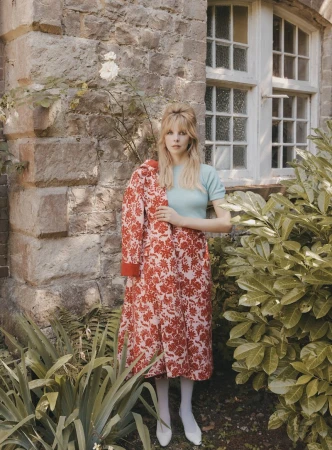 Blonde woman in floral dress poses in garden setting in front of stone building, holding the dress jacket on her shoulder.