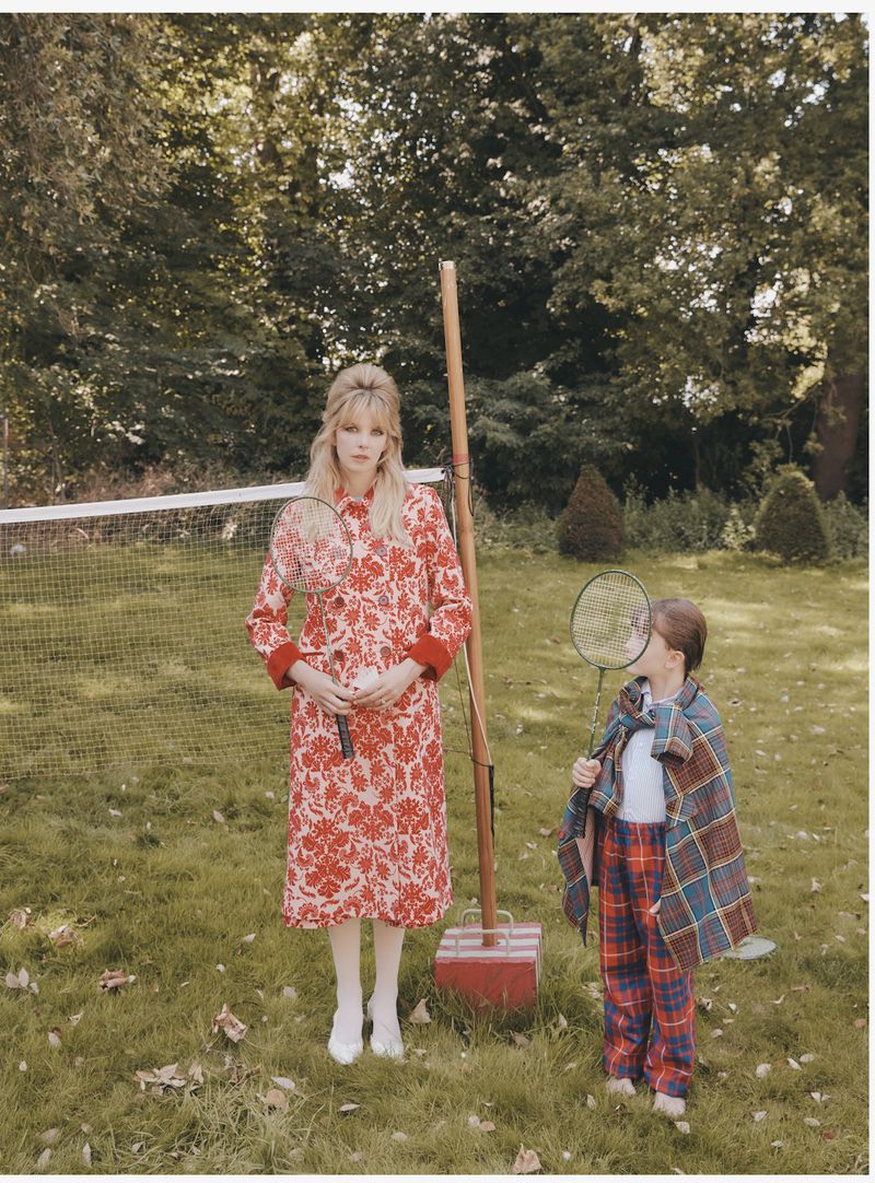Woman and child in floral print fashion, holding tennis rackets on green grass near tennis court and trees in an outdoor setting.