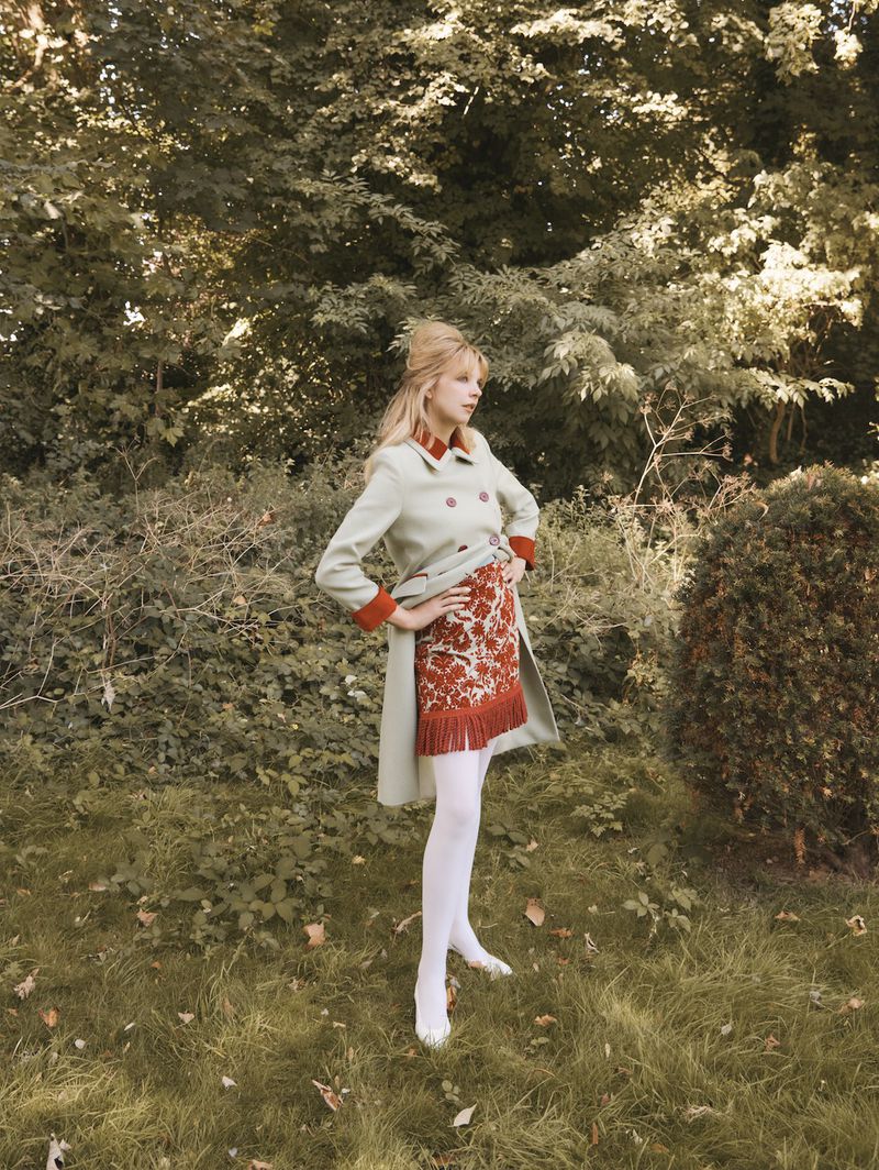 A fashionable young woman stands in an outdoor setting. She wears a light green trench coat and patterned skirt. The backdrop features lush greenery.