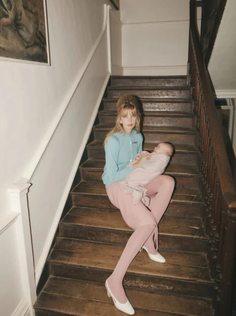 Woman in blue cardigan and pink skirt sits on wooden stairs holding a baby wrapped in a pink blanket indoors with vintage painting.
