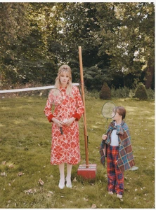 Model wearing a patterned red and white coat holding a badminton racket standing near a badminton net on grass