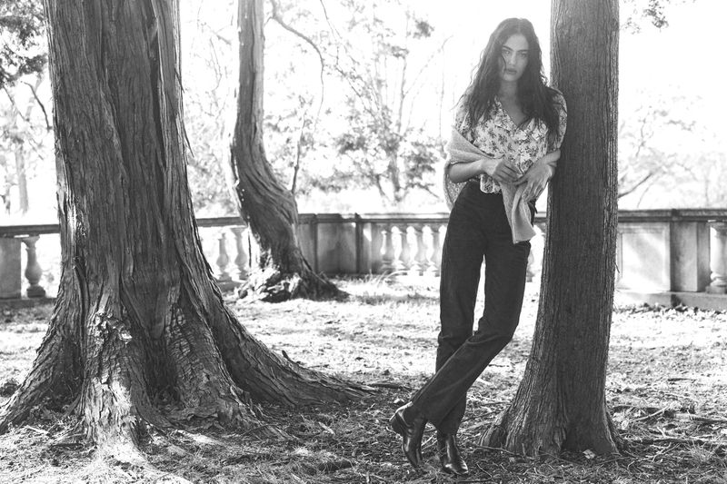 A long-haired woman poses casually in a sun-drenched monochrome outdoor shot. She leans against a tree, creating a natural fashion moment.