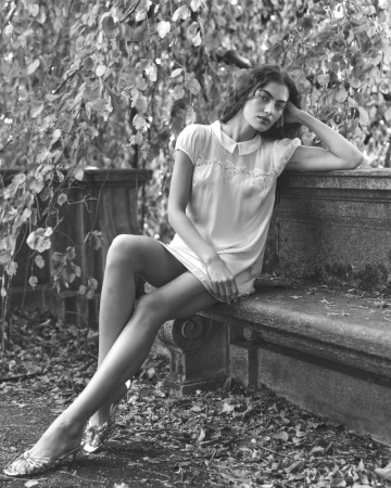 Black and white fashion portrait of a woman in a dress, sitting on a stone bench in an outdoor environment with trees and foliage.