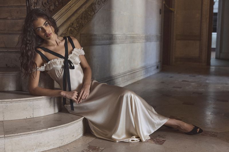 Elegant adult female in a champagne-colored dress with black straps seated on marble stairs inside a grand building with neutral walls.