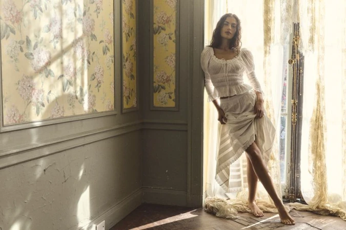 A woman in a white top and skirt poses near a window in a vintage-style room with floral wallpaper and warm, diffused natural light.