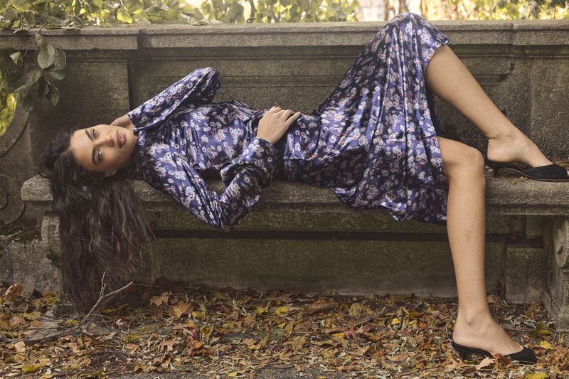 Alana Felisberto reclines on a stone bench wearing a blue floral satin dress and black mule heels in an autumn park setting.