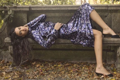 Alana Felisberto reclines on a stone bench wearing a blue floral satin dress and black mule heels in an autumn park setting.