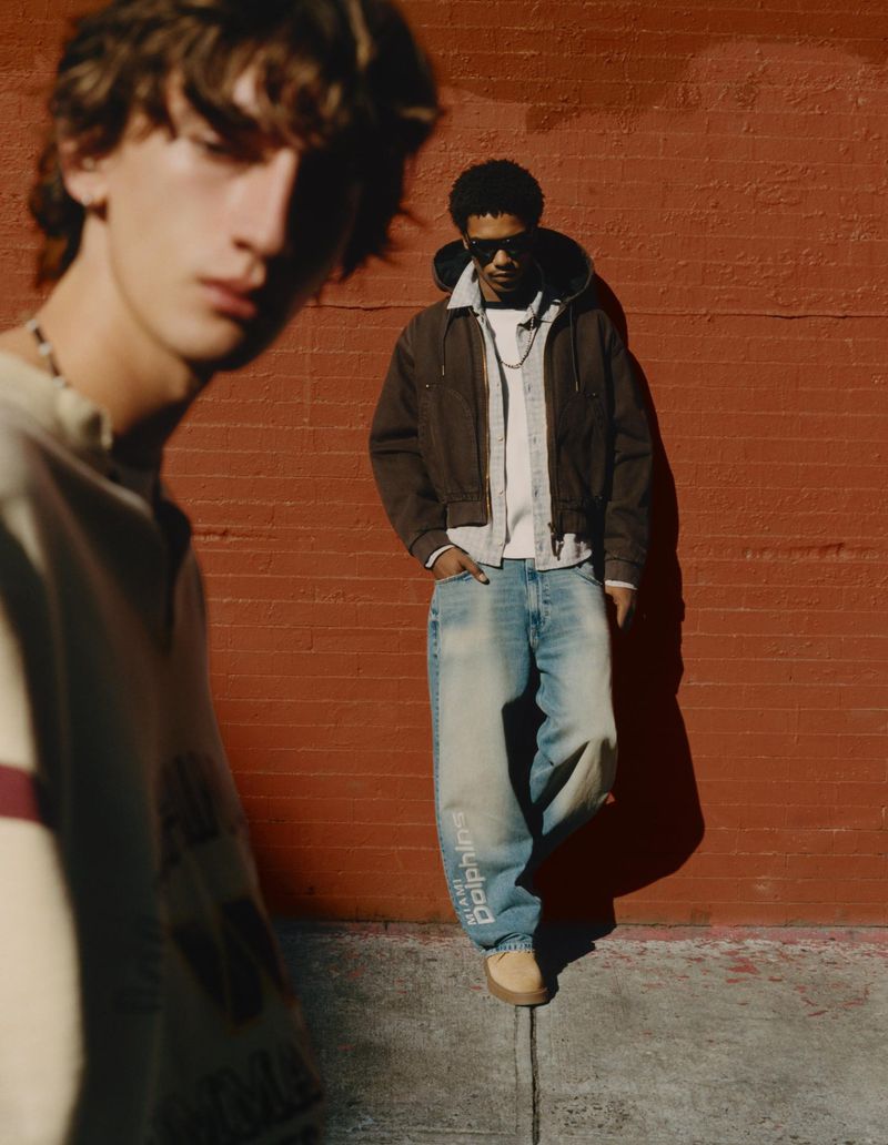 Two young men in casual street style clothing pose outdoors against a brick wall in natural sunlight. One has sunglasses and a jacket.