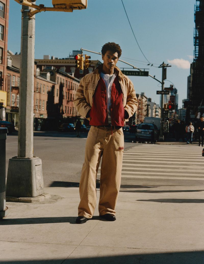 An adult male poses in a tan jacket and pants at a city intersection, with cars, buildings, and traffic lights in the background.