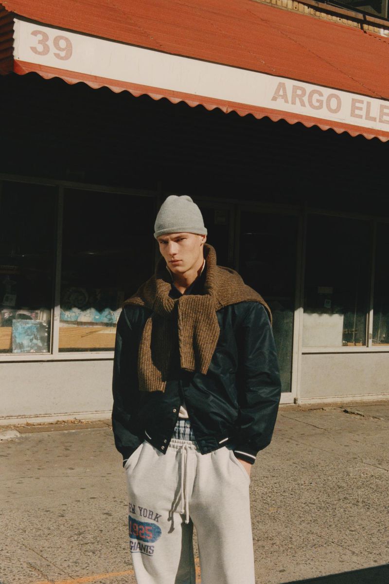 Male model in beanie, scarf, and joggers poses on a city street in front of an urban storefront on a sunny day.