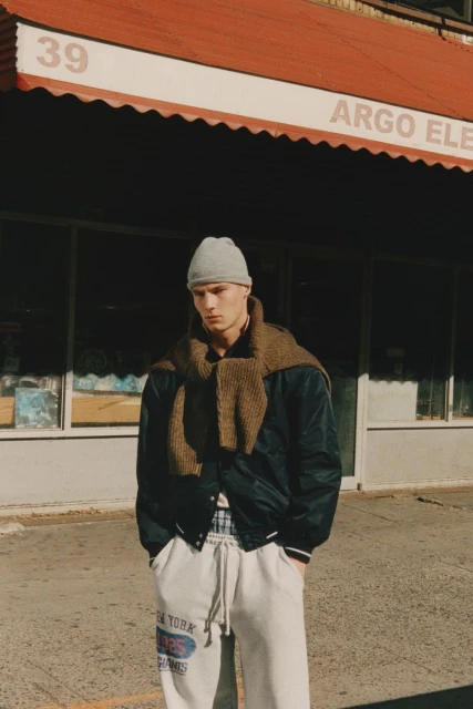 Male model in beanie, scarf, and joggers poses on a city street in front of an urban storefront on a sunny day.