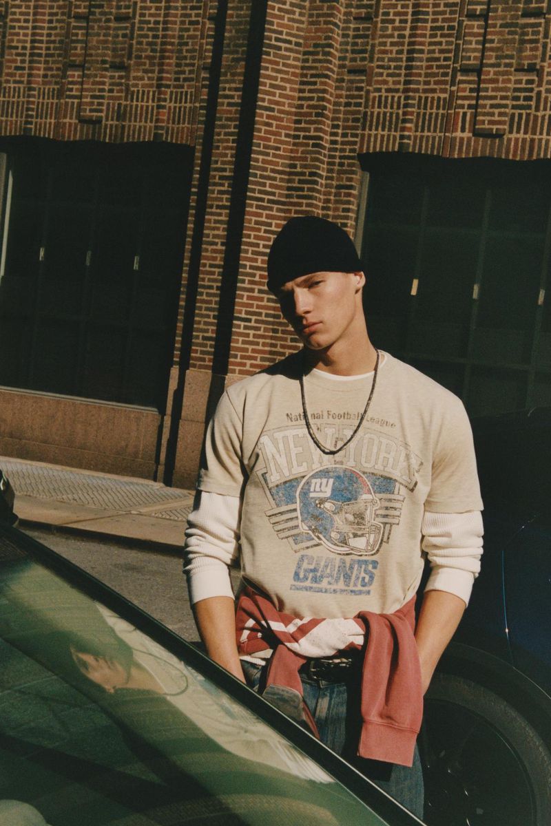 Young man in an urban setting wearing a beanie and football t-shirt poses in front of a brick building and a car.