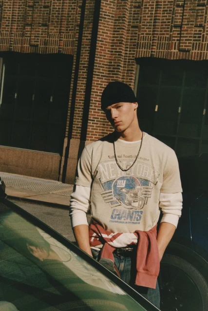 Young man in an urban setting wearing a beanie and football t-shirt poses in front of a brick building and a car.