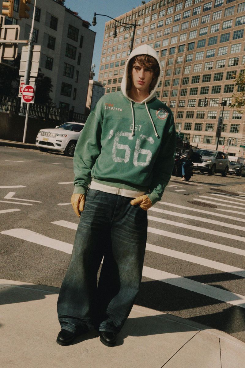 Male fashion model poses on city street in green hoodie, baggy jeans, and black shoes, buildings and traffic in background on sunny day.