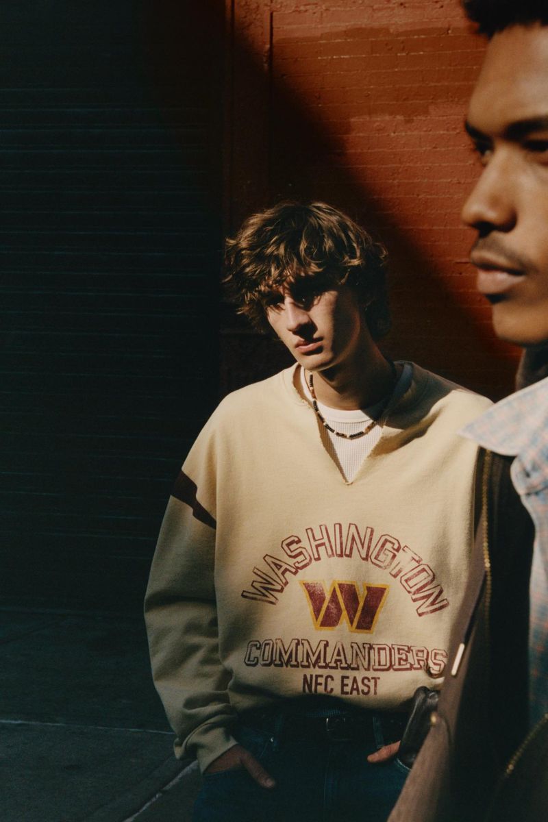 Two young adult men pose in urban setting. One wears a vintage-style cream Washington Commanders sweatshirt and the other a plaid shirt.