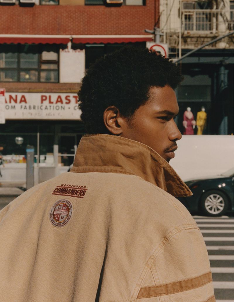 Adult male in urban street setting wears a tan jacket with a sports team emblem, shot from the back in editorial style.
