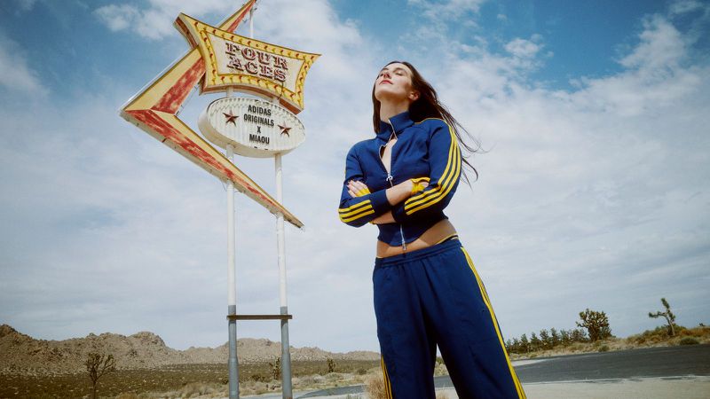 A young adult woman poses in a blue tracksuit with yellow stripes near the Four Aces sign in the desert against a blue sky.