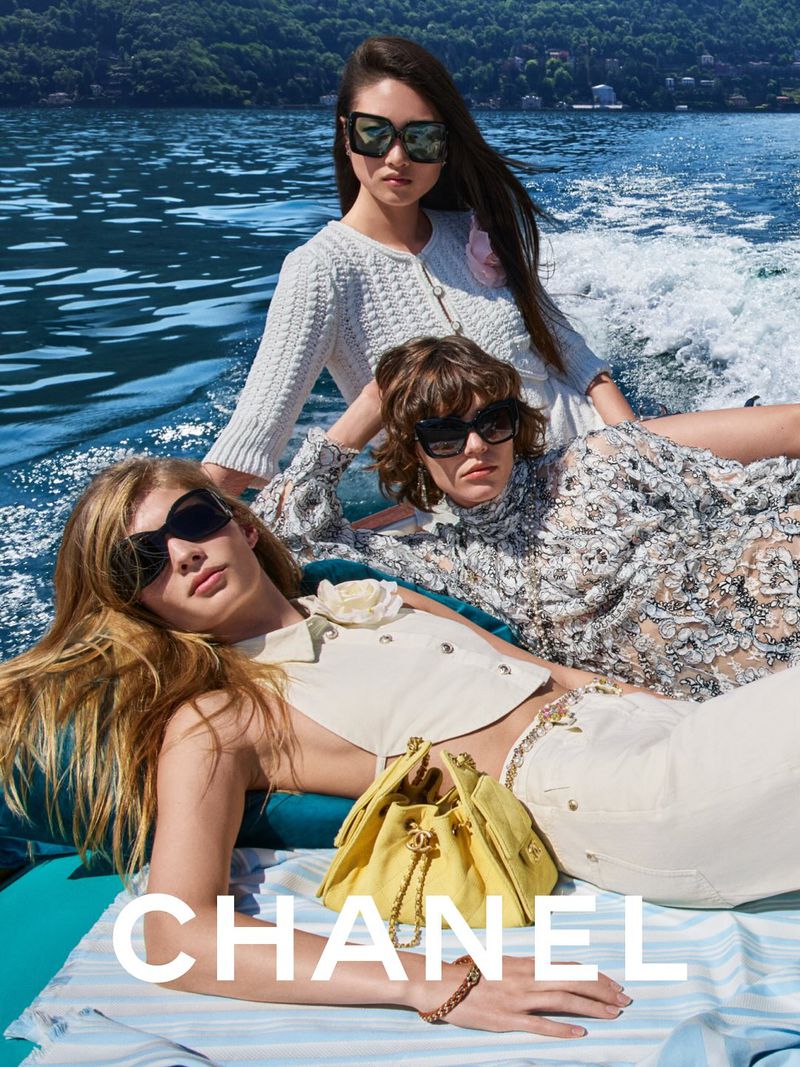 Three fashion models pose on a boat, wearing sunglasses and light-colored garments, with a yellow purse and water in the background.