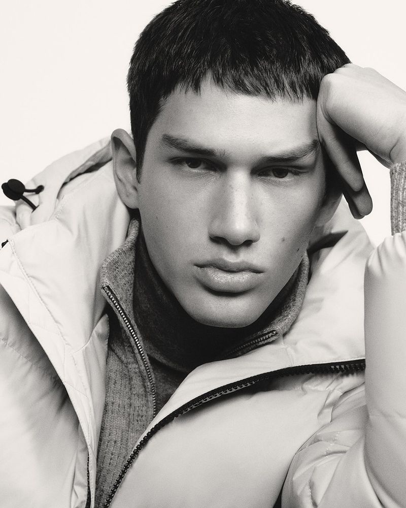 Close-up studio shot of an adult male wearing a puffer jacket and turtleneck sweater with a hand resting on his head.