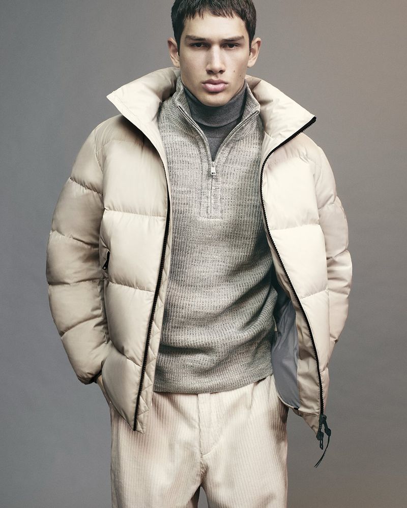 Fashion shot of a young adult male posing in a beige puffer jacket and corduroy pants against a neutral studio backdrop. Modern menswear.