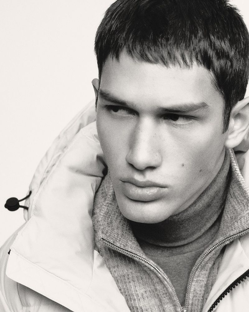 Monochrome portrait of a male model wearing a puffer jacket and turtleneck sweater against a bright, neutral backdrop in a studio shot.