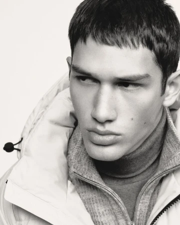 Monochrome portrait of a male model wearing a puffer jacket and turtleneck sweater against a bright, neutral backdrop in a studio shot.