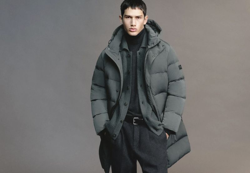 Man in a hooded puffer coat, dark turtleneck, and grey wool trousers stands in a fashion pose against a grey studio backdrop.