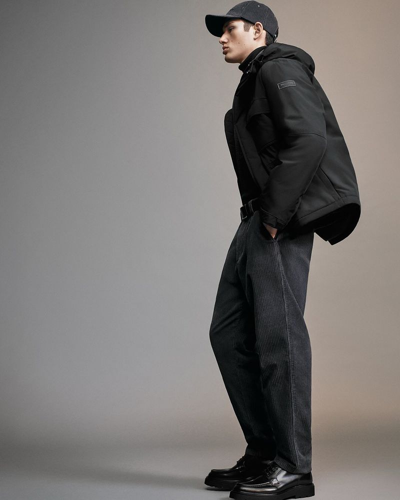 Full-length shot of a young adult male wearing a black jacket, dark grey pants, black cap, and black shoes, studio lighting on a grey background.