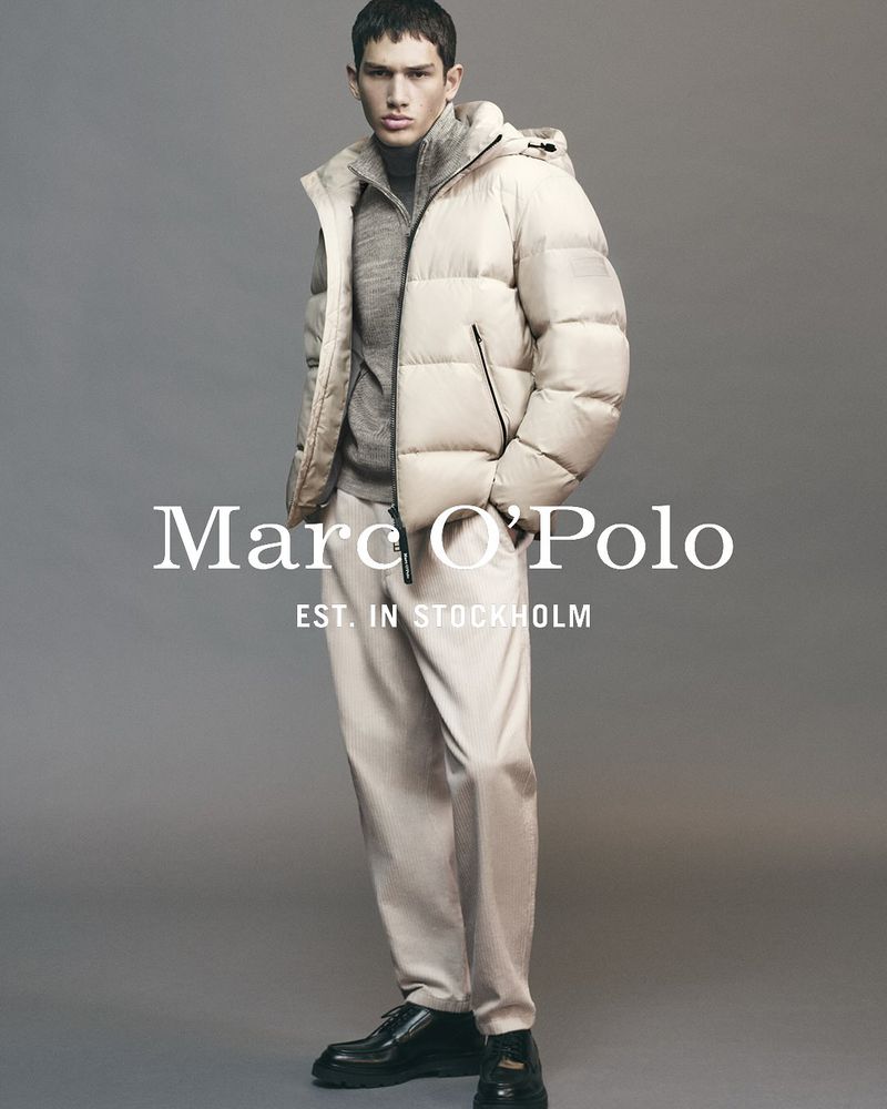 Man posing in a down jacket and trousers against a grey backdrop, showcasing a minimalist aesthetic with the brand logo.