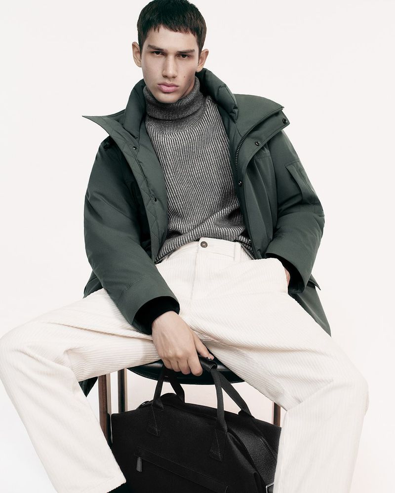 Adult male fashion model poses while seated in an olive down coat, gray turtleneck sweater, and white corduroy pants with a black bag.