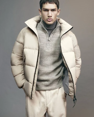 Model Luka Tesic posing in a beige puffer jacket, grey half-zip knit sweater, and light corduroy trousers against a studio background.