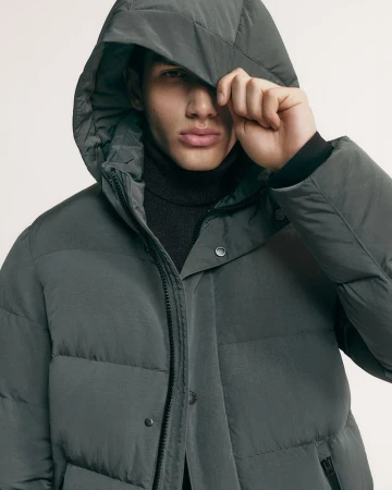 Model Luka Tesic wearing a grey hooded puffer jacket and black turtleneck sweater against an off-white studio background.