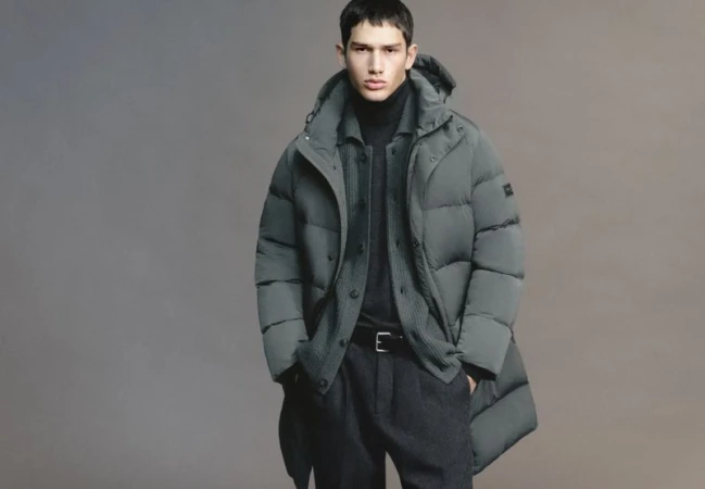 Model Luka Tesic wearing a grey hooded puffer jacket layered over a knit cardigan and black trousers against a studio backdrop.