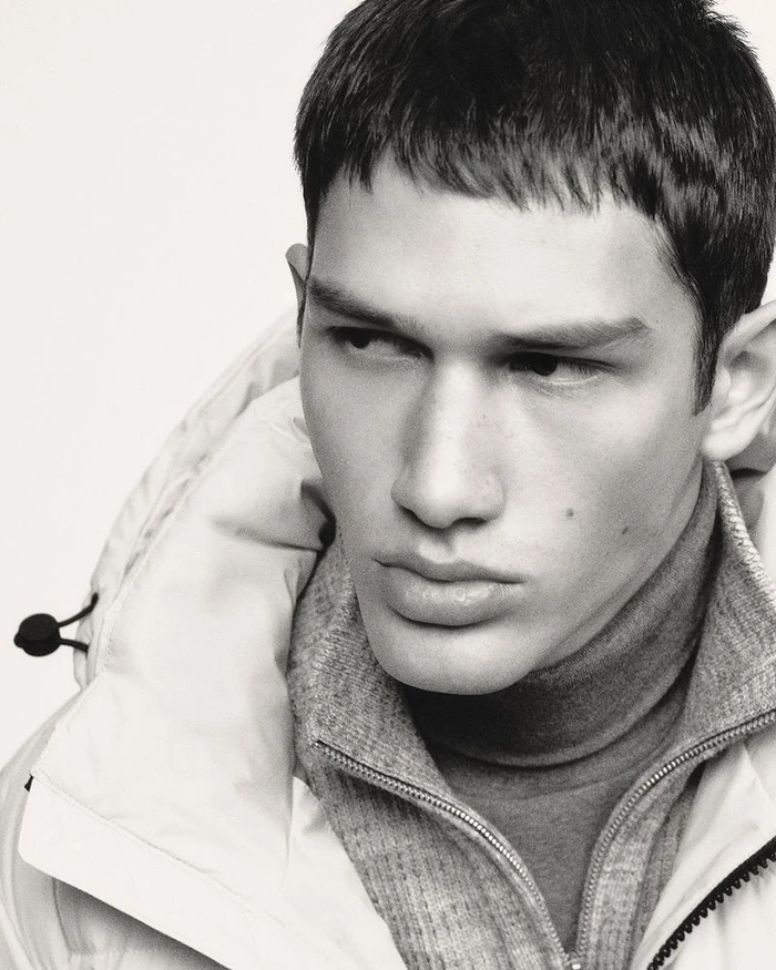 Close-up black and white portrait of model Luka Tesic wearing a layered puffer jacket and knit collar against a seamless backdrop.