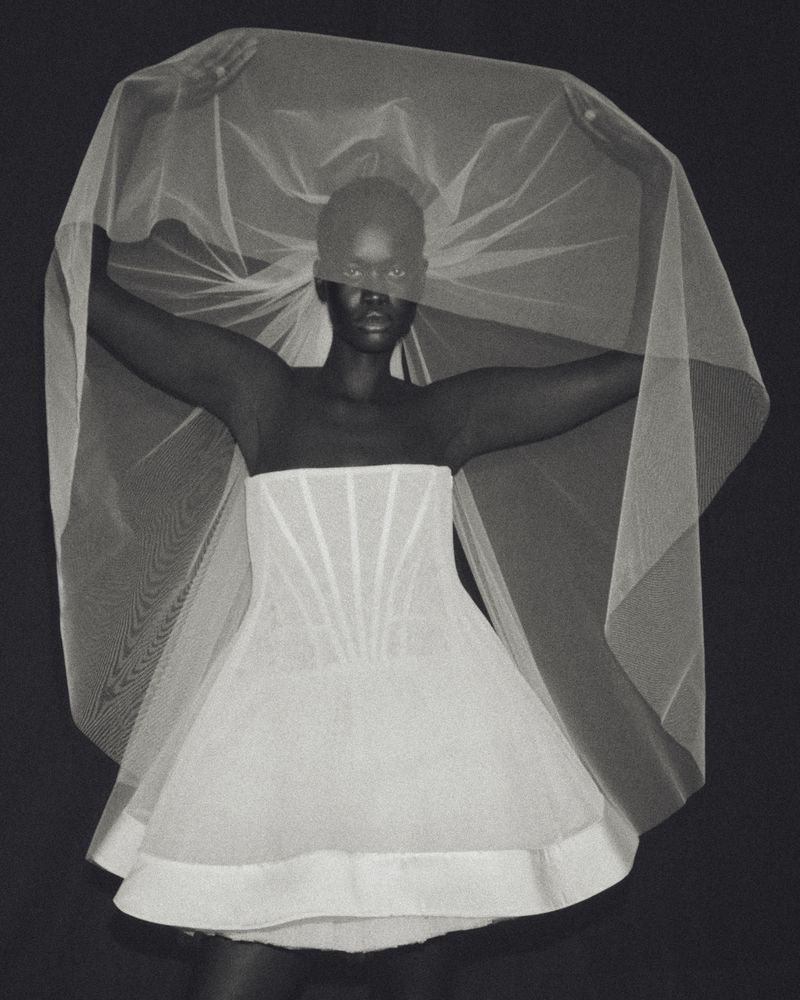Black model wearing a strapless white corset mini dress and holding an expansive sheer bridal veil against a black background.