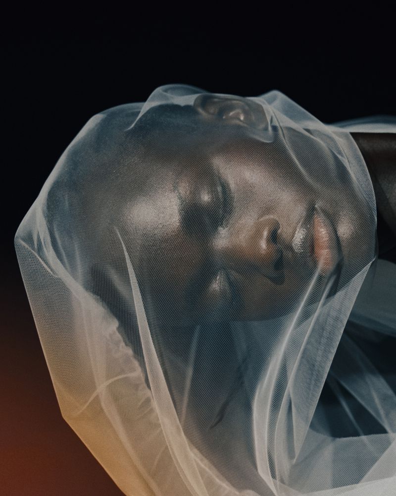 Close-up of a woman's face covered with sheer fabric against a dark background in a studio setting, showcasing beauty and fashion.