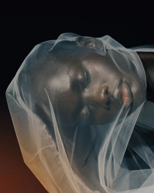 Close-up of a woman's face covered with sheer fabric against a dark background in a studio setting, showcasing beauty and fashion.
