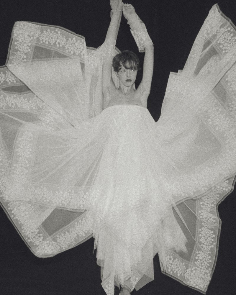 Model posing with arms raised in an expansive white tulle bridal dress with lace embroidery against a solid black background.