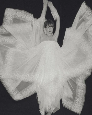 Model posing with arms raised in an expansive white tulle bridal dress with lace embroidery against a solid black background.