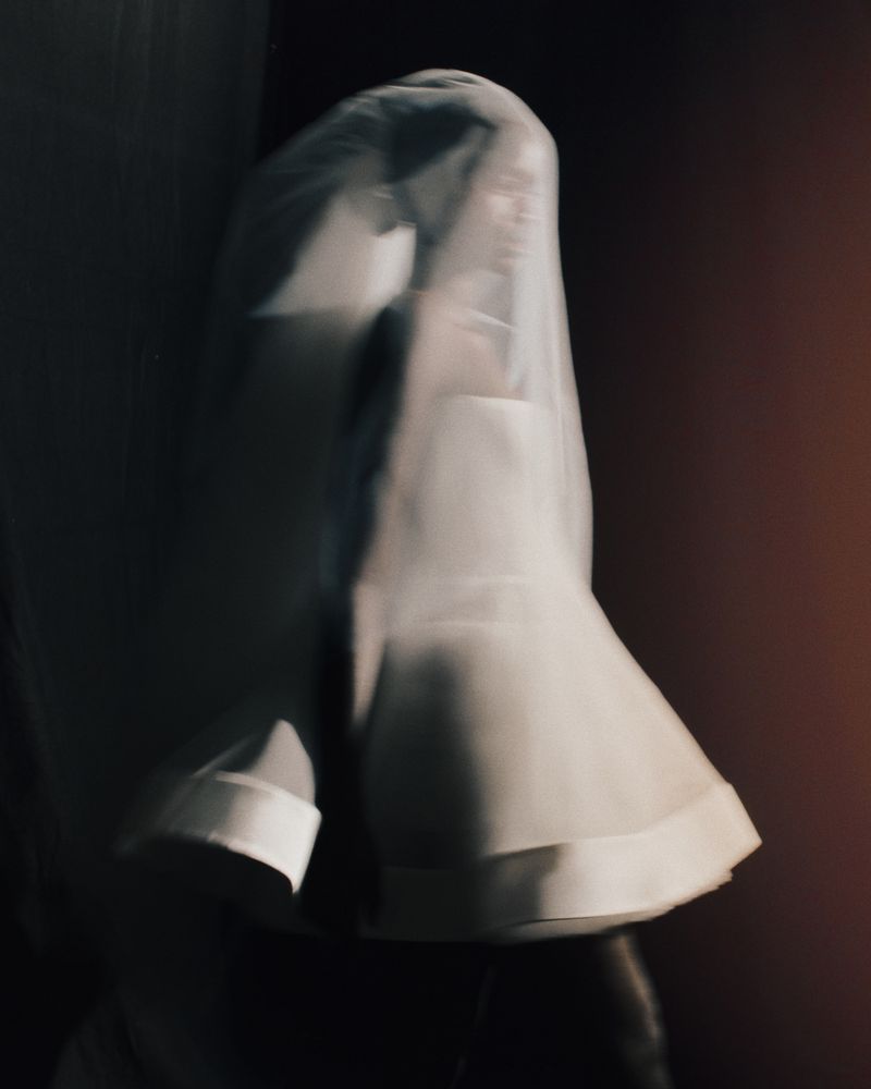 Person covered in a sheer white veil standing in a dark room with a reddish-brown wall, soft diffused lighting, and a blurred effect.