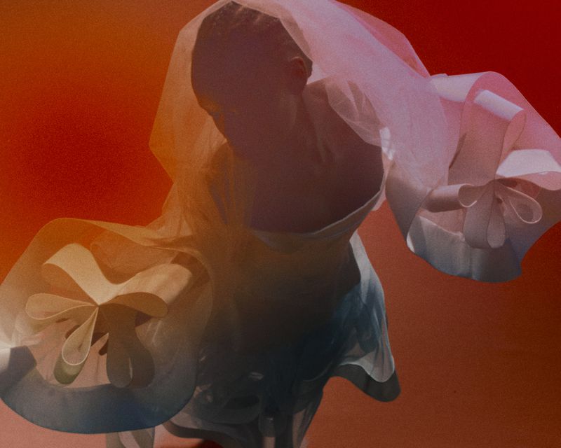 Woman in flowing white top and sheer cape against gradient red background, soft lighting, abstract fashion portrait.