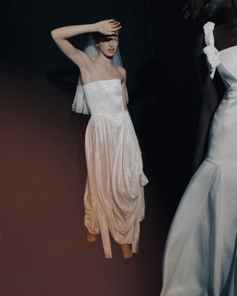 Two women pose wearing wedding dresses against a dark backdrop in a studio setting, illuminated by focused lighting to highlight their attire.