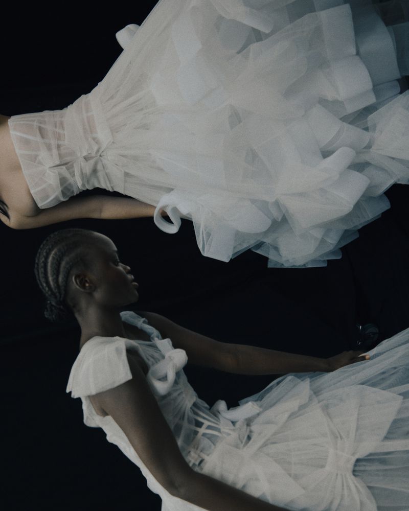 Two women wearing white tulle dresses pose against a black background, one laying down and the other sideways for a fashion shot.