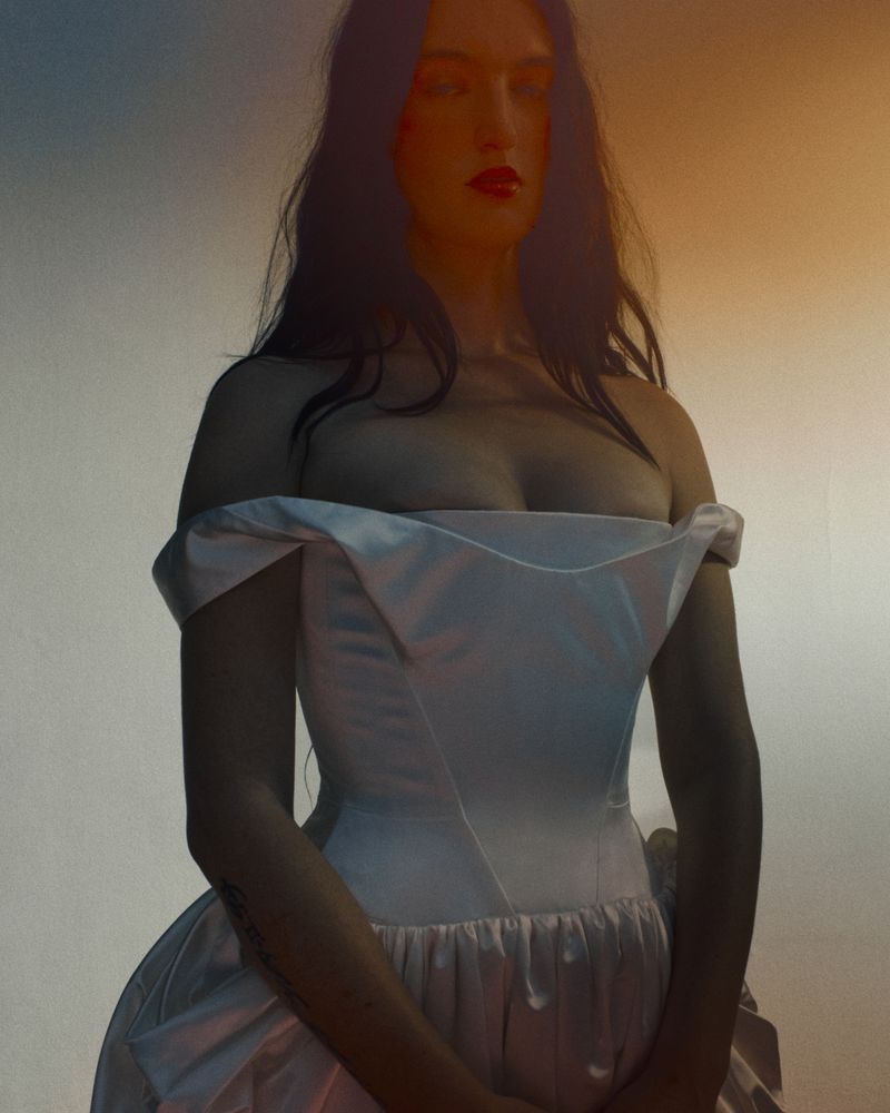 A woman with dark hair, wearing a white dress and red lipstick, poses in a medium shot against a soft gradient background.