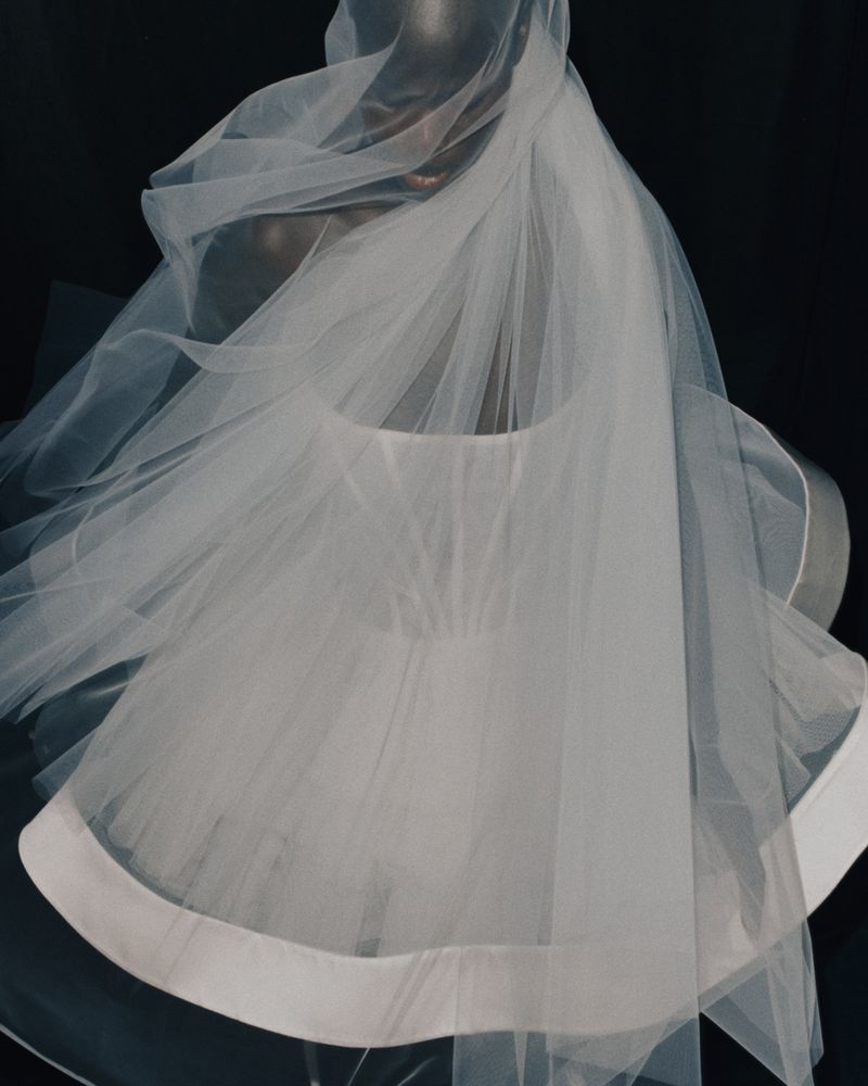Close-up shot of a mannequin draped in a flowing white tulle veil, with a black backdrop creating a dramatic studio effect.