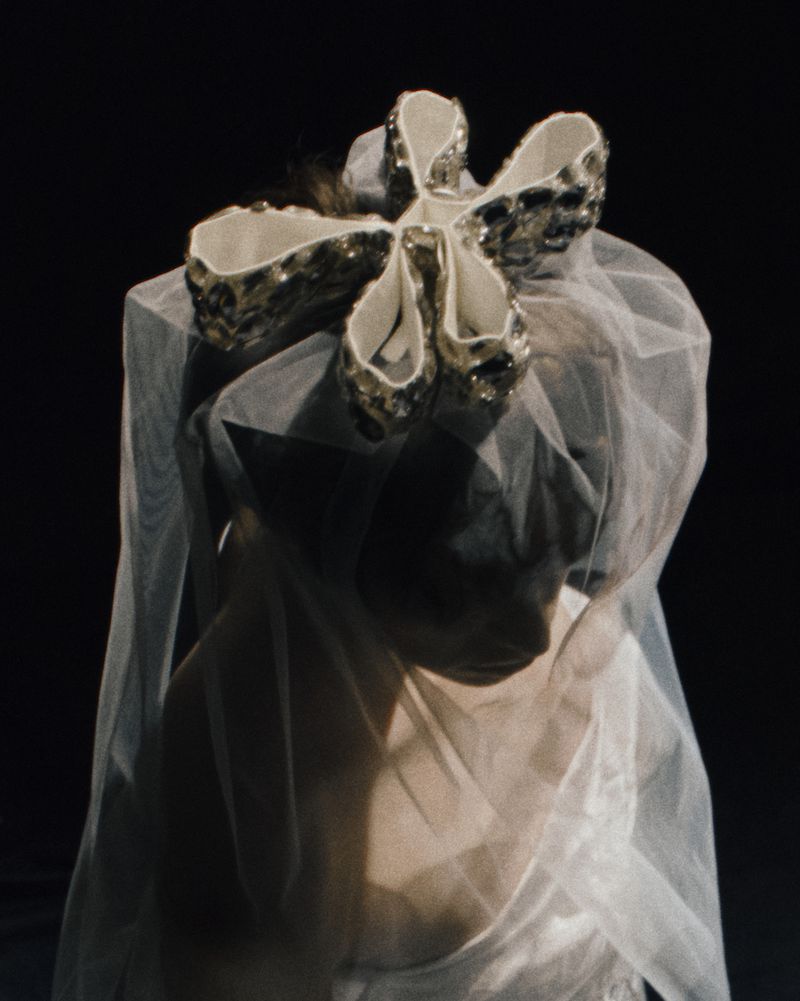 Woman with sheer white veil and large decorative flower on head, against dark background. Close-up captures ethereal fashion aesthetic.