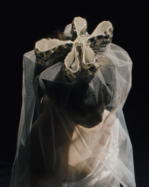 Woman with sheer white veil and large decorative flower on head, against dark background. Close-up captures ethereal fashion aesthetic.