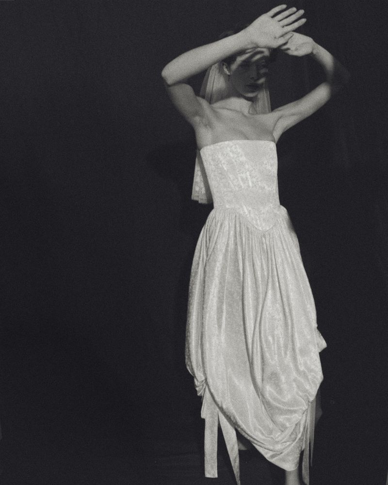 Monochromatic portrait of a fashion model with a white gown, covering her face with her hands in front of a black studio background.