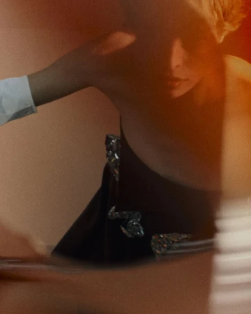 Model with blonde hair poses in front of an orange background, wearing a black dress with shiny embellishments and a white sleeve.